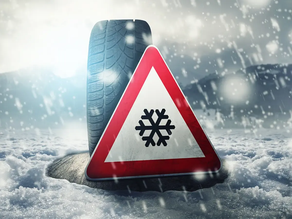 Verkehrszeichen mit Schneeflockensymbol vor einem Winterreifen in einer verschneiten Winterlandschaft.