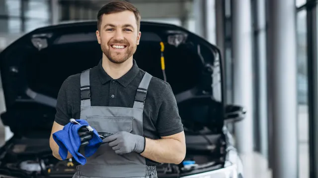 Lächelnder Kfz-Mechaniker in Arbeitskleidung hält Werkzeug und Tuch, im Hintergrund Auto mit geöffneter Motorhaube
