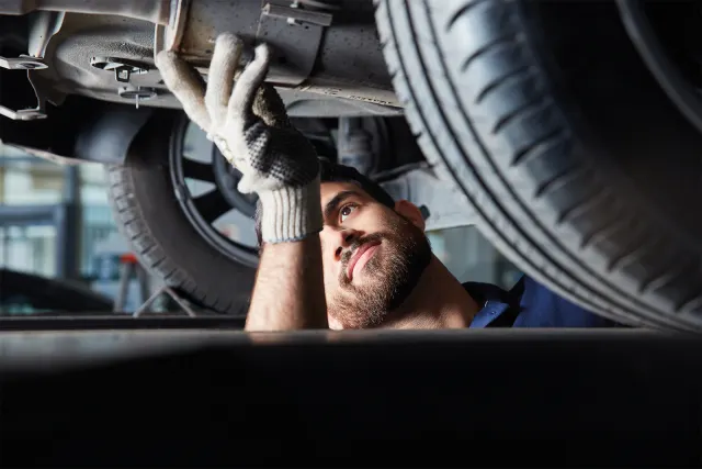 Mechaniker mit Handschuhen prüft Unterseite eines Autos, Reifen im Vordergrund