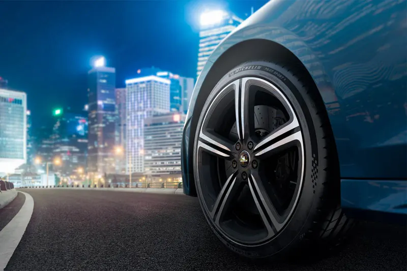 Vorderrad mit Michelin Reifen an einem blauen Auto, Stadtlichter und beleuchtete Hochhäuser bei Nacht unscharf im Hintergrund