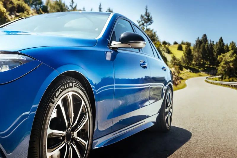 Seitenansicht eines blauen Autos auf kurviger Landstraße bei sonnigem Wetter, Michelin Schriftzug am Vorderreifen sichtbar