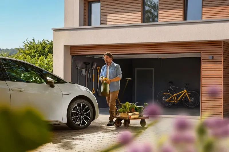 Mann mit Gießkanne vor einer Garage neben weißem Auto, Fahrrad und Gartenwagen.