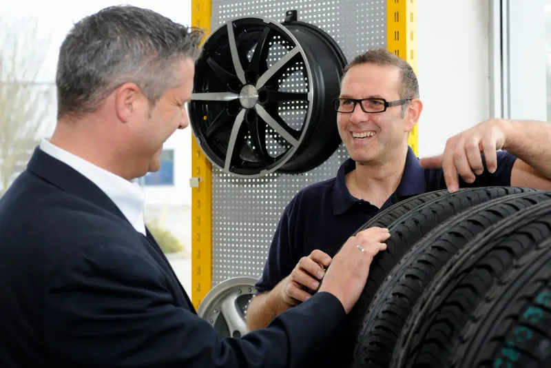 Bild Verkäufer berät Kunden zu passenden Autoreifen im Verkaufsgespräch, im Hintergrund ist ein Reifenfelgen zu sehen